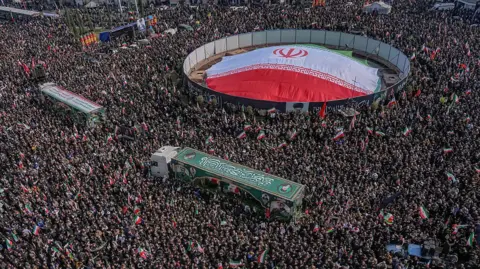 Getty Images People wave Iranian national flags as they attend funerals for commanders from the Islamic Revolutionary Guard Corps (IRGC), the army and others killed in the early days of U.S. and Israeli strikes on Iran at Enghelab (Revolution) Square in Tehran.