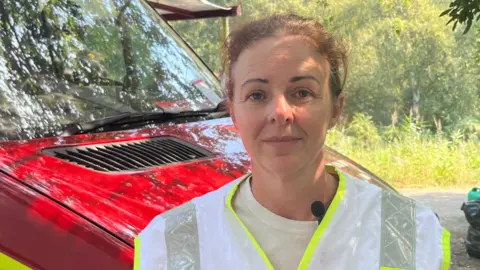John Devine/BBC Vicky Best is looking straight at the camera with a neutral expression. She has red hair in a ponytail and is wearing a white T-shirt and a yellow and silver Hi-Vis jacket. She is standing in front of a grassy area and a red and yellow response vehicle.