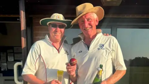 Nic Downes Nick Downes holding up the ball he took his 1000th wicket for whilst holding a bottle of champagne. He is pictured with the man he got out - both are in hats and cricket whites.