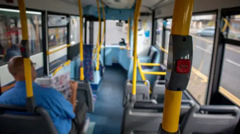 Getty Images A man sits in an empty bus reading a newspaper. He is wearing a blue shirt and glasses. The bus is travelling on a road. 