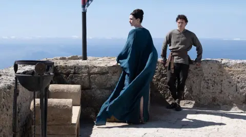 Universal Anne Hathaway is wearing a flowing blue dress and Tom Holland a brown shirt and dark leggings. The actors are standing in sunshine at the battlements of a castle.