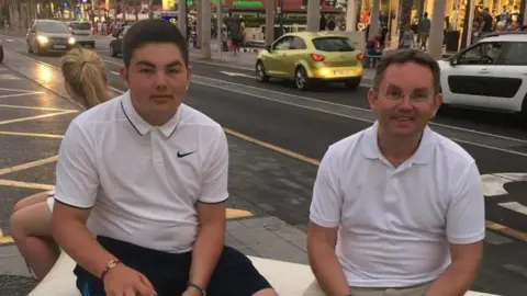 Family handout Andrew Rowlands and his father John. They are sat next to each other on a bench by a busy shopping street. Andrew has black short hair and is wearing a white Nike branded polo and black shorts. Andrew has light brown hair and glasses and wears a white polo and beige trousers. They are both slightly smiling at the camera.
