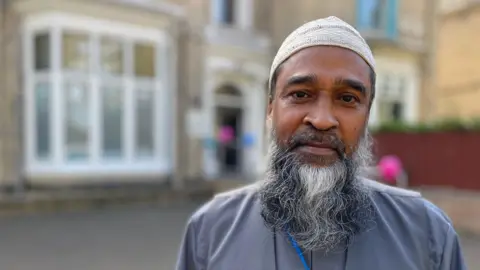 Raj Choudhury stands outside the mosque in Pearson Park, Hull. He wears a white skullcap and has a black and white beard.