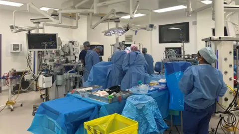Royal Papworth Hospital An operating theatre at Royal Papworth Hospital in Cambridgeshire. Six staff with blue scrubs and plastic gowns are performing an operation. In the foreground is a nurse looking at a table where equipment is being prepared. This includes metal surgical instruments. In the centre, five staff are gathered around a patient on an operating table. The theatre is modern and has computers and lighting attached to the ceiling.