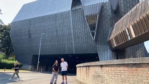 Two people are standing in front of a large, metal elephant-shaped building with a road running underneath it.