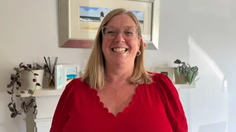 Susan Terris has straight, shoulder-length, blonde hair. She is smiling and wearing large clear-framed glasses. One hoop earring can be seen on her right ear as her hair is tucked behind it. She is also wearing a delicate silver necklace and a red top with ruffled edge around the neck. She is standing in front of a white wall which is decorated with a number of paintings and plants sitting on a fireplace mantlepiece.
