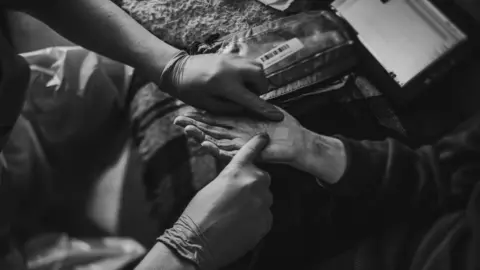 SCAS Black and white image of a a pair of hands. One is wearing gloves and the other has a plaster being placed on her palm. There is medical kit in the background.