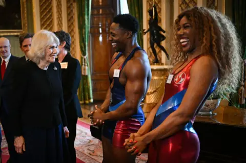 Justin Tallis/PA Media Queen Camilla, wearing a black dress, smiles as she meets Gladiators Nitro and Fire, who are dressed in their gladiator outfits and smiling back at her, in a grand room of Buckingham Palace