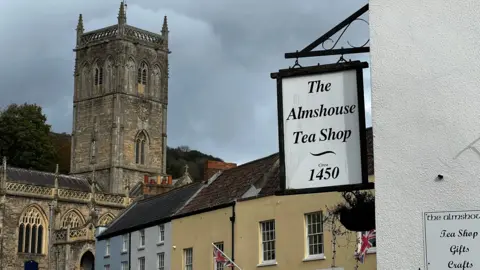 Una vista de la plaza de la ciudad de Axbridge, hacia la antigua iglesia. En primer plano, un cartel dice "La tienda de té Alms House, fundada en 1450."