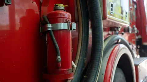 Getty Images A close-up of the side of a fire engine. The red vehicle's black water hose can be seen, just in front of a wheel arch.