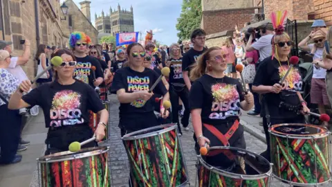 A march through one of Durham's cobbled streets during the Pride celebrations. Women wearing shirts with the words "disco queen" and "disco fever" are beating colourful drums. The people in the crowd wear rainbow-coloured attire, including rainbow afro-style wigs. Some don colourful face paint. In the background, a trade union's banner can be seen waving, and in the distance is Durham Cathedral.