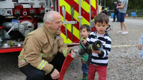 A firefighter with short grey hair wearing his uniform and kneeling down beside a young boy, who is wearing a black and white striped shirt. They are beside a fire engine and are playing with a water hose which is used to put out fires.