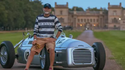 Ian Jones, dressed in a stripey T-shirt, brown shorts and a blue cap, rests on the front right wheel of his RA4 Vanguard car, which is park on the driveway of a large house