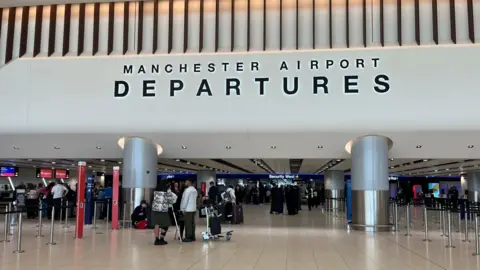 Manchester Airport departures lounge - with security west sign straight ahead. Some passengers queue with luggage.