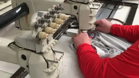 AFCF / FREEDOM OF INFORMATON A person operating an embroidery machine.  They are wearing a top with red sleeves feeding a grey top under a stitching machine.