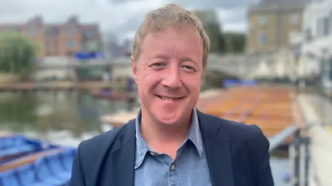 PHIL SHEPKA/BBC Mayor Paul Bristow smiling at the camera. He is wearing a navy blazer and a denim shirt. In the background is the River Cam, out of focus. 