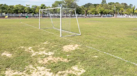 A white football net on a pitch with large patches of bald and dry grass. A fence runs along the edge of the pitch in the background with trees behind.