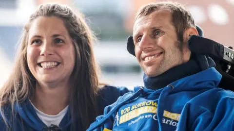 Lindsey Burrow, who has long, dark brown hair and is wearing a blue hooded top. She is smiling and kneeling down next to Rob Burrow who is in a wheelchair which has a neck support. He is smiling and is also wearing a blue hooded top with yellow writing on which says 'Rob Burrow Leeds Marathon'.