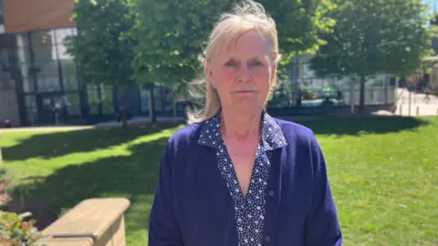 BBC/Mark Ansell Christine standing outdoors in a landscaped area on a bright, sunny day. She is positioned centrally in the frame and faces the camera directly, standing on a paved area with grass and trees behind them. She is wearing a dark blue cardigan over a patterned blouse, with light‑coloured hair pulled back and small earrings visible.