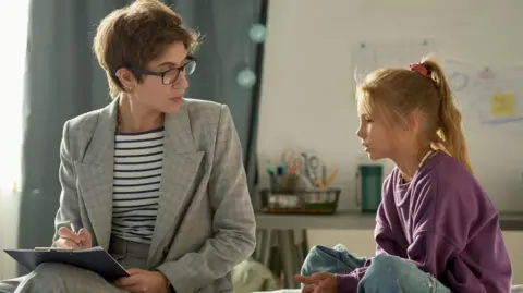 A young girl talking to social worker about her problems while they sitting in bedroom at home - Stock Shot. The girl has a blonde ponytail and is wearing jeans and a purple top. A woman wearing a grey suit and holding a pen and clipboard is sitting next to her on the bed.