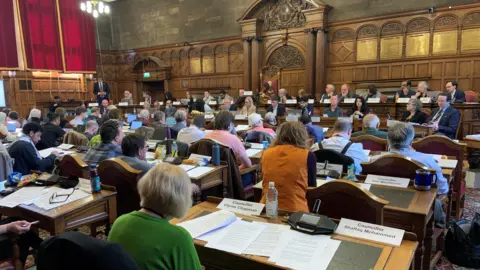 Sheffield council chamber has rows of desks and chairs with dozens of councillors. They are facing a row of senior councillors, officers and the Lord Mayor sitting in a large chair in robes and chains