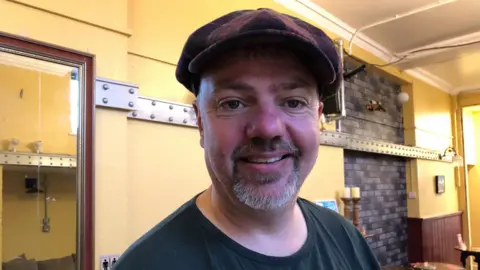  Darren Rozier/BBC A man with a goatee beard and a flat cap is smiling at the camera. He is wearing a black T-shirt and is standing indoors in a cafe with cream-coloured walls. 