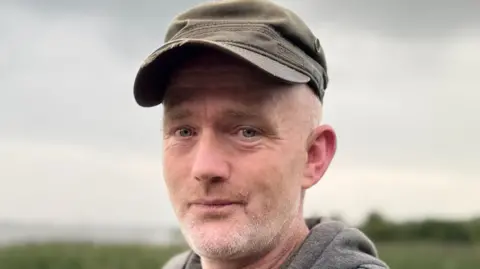 A middle aged white man is wearing a grey hoodie and a brown peaked cap. He has some white stubble on his face. 