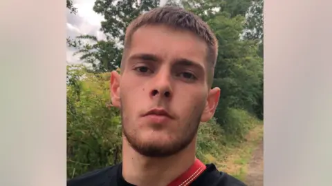 Joshua Stewart - a man in his early 20s with brown hair, dark eyes and a short beard. He is looking at the camera and there are trees and bushes in the background. He is wearing a black top.