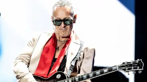 Getty Images Vivian Campbell está no palco. Ele está tocando um violão. Ele está usando óculos de sol e uma jaqueta prateada com um lenço e colares vermelhos. Branco e azul estão brilhando atrás dele de uma tela grande.