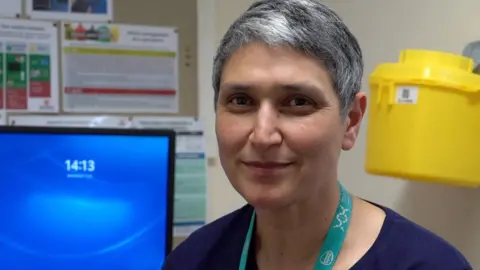 A woman in a lanyard with short grey hair smiles while sitting by a desk in a hospital.