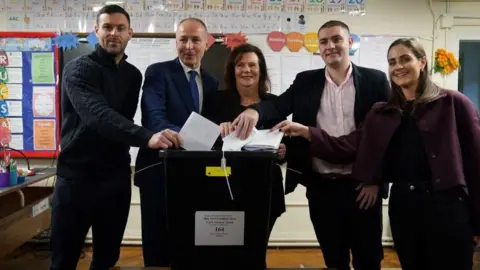 PA Media Micheál Martin stands with his family in a primary school classroom as the members place their white ballot papers into a black vote box. 