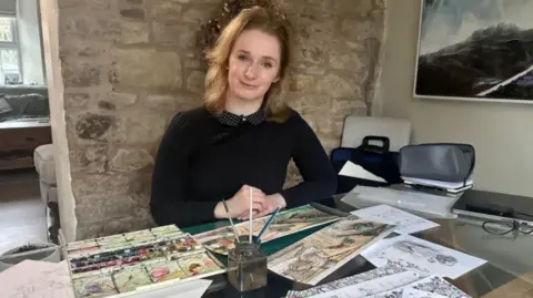 A woman with tied back blonde hair and wearing a black jumper sits in front of a table with paint and brushes and sketches
