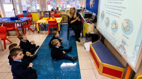 PA Media Children sit below an interactive whiteboard in a colourful classroom. A woman with blonde hair is teaching them.