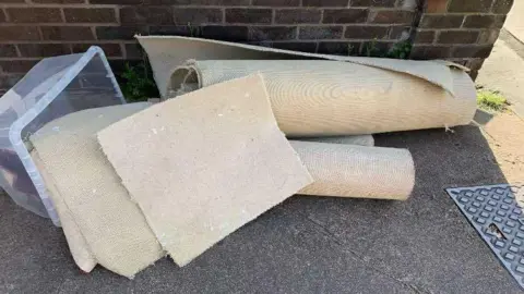 Sea Adora White rugs rolled up on the pavement. There is a plastic box on its side with off-cuts of carpet in it.