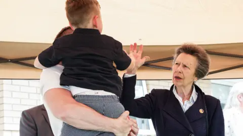 Press Eye The Princess Royal, a woman with grey hair, wearing a blue coat, puts her hand up to a boy in a black top, being held by a man with a white top.