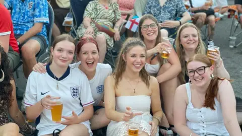 A group of young women holding pints of beer are sat in a group. Some are wearing England shirts.