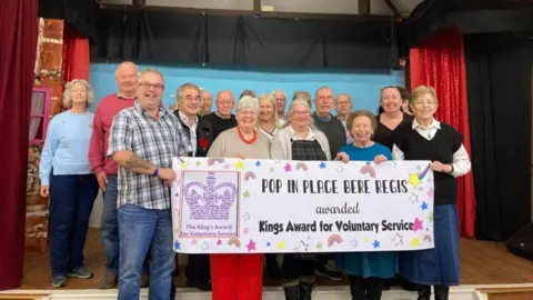 A crowd of people from Pop in Place Bere Regis stood on a stage holding a large banner which says "Pop in Place Bere Regis awarded King's Award for Voluntary Service"