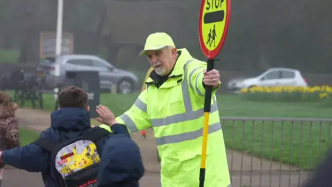 A lollipop man in a yellow hi-vis jacket does a high five with a pupil who is running past.
