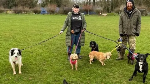 Lizzy's Pet Services Two people, in a field, out walking seven dogs. Both people are holding leads, looking at the camera and smiling. Lizzy has on a long green coat, dark trousers, black top and has black and white hair. Tom has on a black hat, green coat, trousers boots and a top. He has a ginger beard. Trees are behind them. 