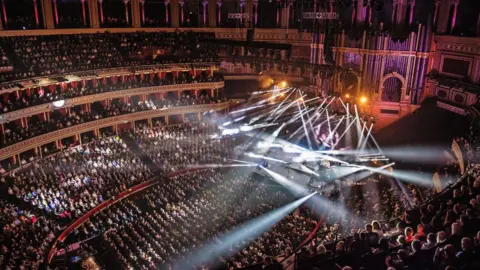 Getty Images View of a packed audience inside the Royal Albert Hall during a live concert, with dramatic stage lighting and beams shining out into the crowd.