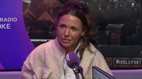 A woman with long dark hair, wearing a beige fluffy jacket over a white hooded top, sits and smiles in front of a purple BBC Radio Stoke microphone.