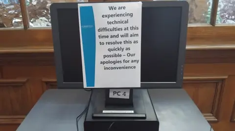 Contributed A black desktop PC screen in an Edinburgh library that has been switched off and has an A4 message stuck to it. The notice reads: "We are currently experiencing technical issues and will aim to resolve this as quickly as possible. We apologize for any inconvenience caused". The screen is placed on a gray table in front of a wooden background with frosted glass windows.