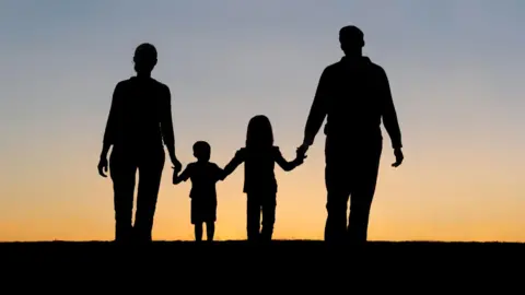 A man and woman holding hands with their son and daughter. They are silhouetted in the photo with the sunset behind them.