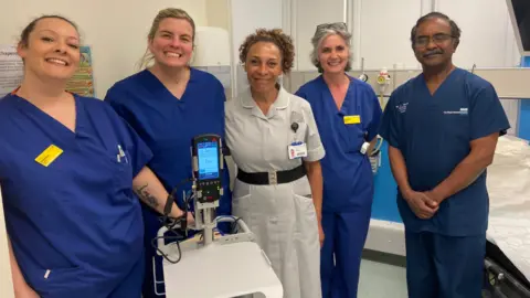 Four women and one man stand by the new machine on a ward in the hospital. They are in blue scrubs with one women in a grey nurses uniform.