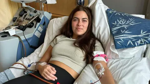 Molly Morin Molly is smiling at the camera as she lies down in her hospital bed. There are tubes going into her arms and there's blood flowing through the tubes. There's hospital machinery to her left and her right arm is lying straight on the bed while her left arm is lying over her waist.