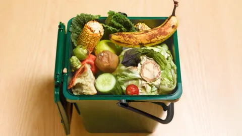 Discarded food in a green waste bin on a wooden floor in a kitchen.