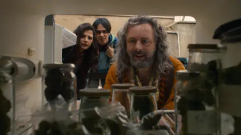 Out There A man with long, curly grey hair and facial hair, staring into a cupboard full of jars. A young man and young woman, both brunette, are stood behind him, also leaning down to look at what he is looking at. They have puzzled looks on their faces.