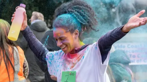 Caroline James Photography A woman in a park with long brown hair and wearing a white t-shirt has finished a run as part of the Holi festival of colour. She has both arms reaching out and is holding a multi-coloured water bottle in her right hand. A rainbow of powered colour is over her face and t-shirt. She is smiling.