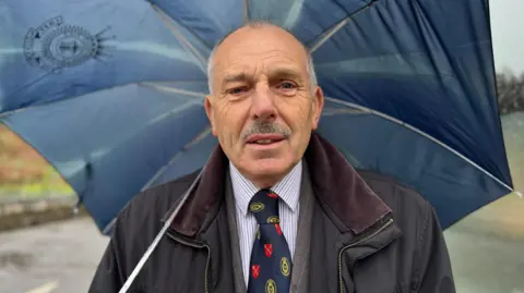 Andrew Smith is wearing a suit and tie under a dark jacket and is holding a dark blue umbrella. He has some grey hair on either side of his head and a moustache. 