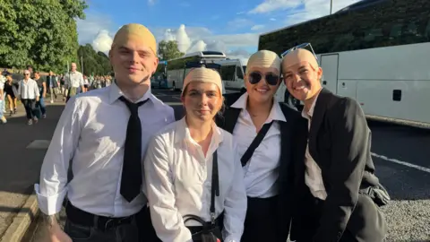 A man and three women. They are all wearing bald caps. The two women to the right are wearing black jackets.